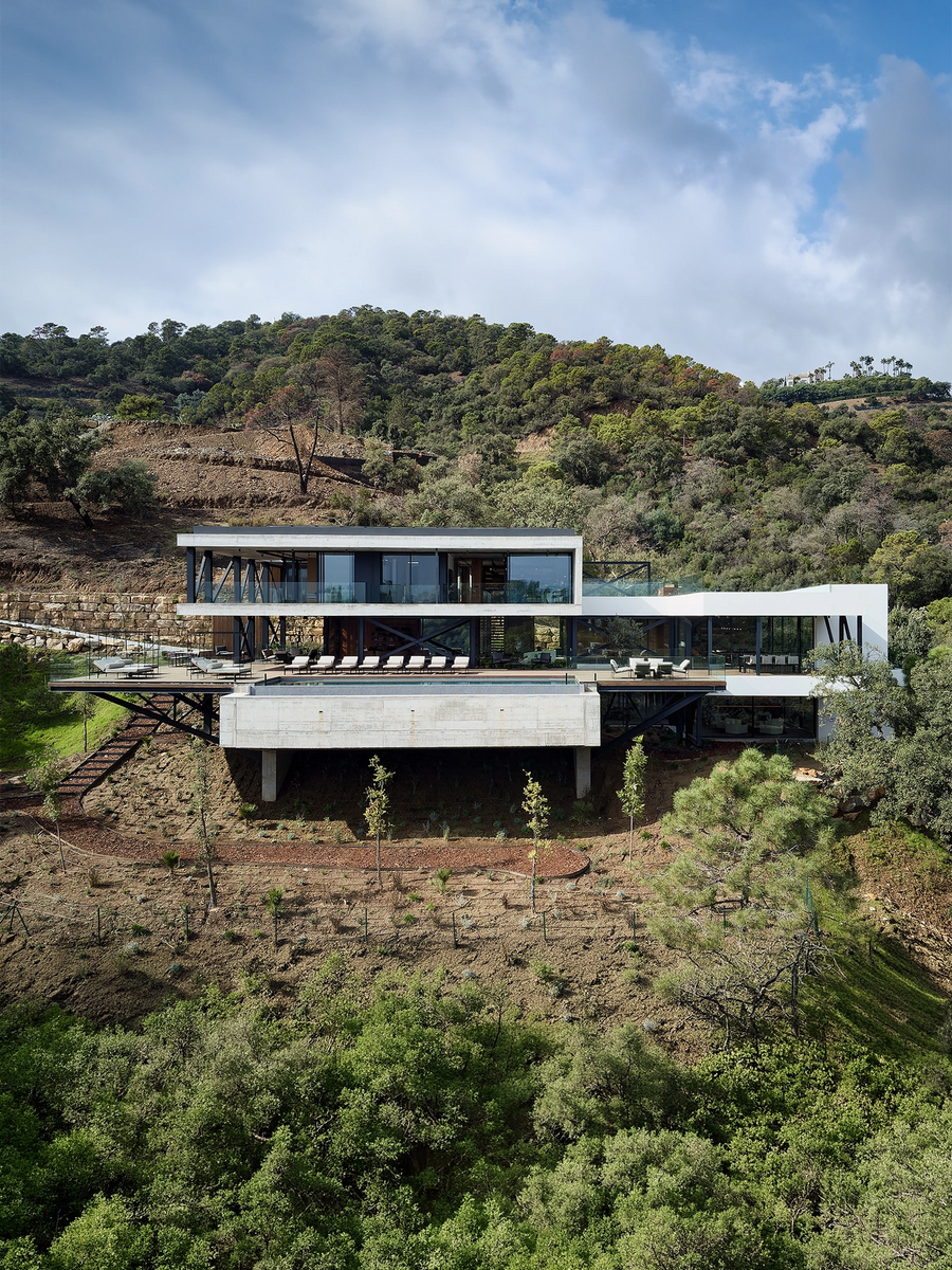 Modern elevated concrete house with large glass walls, set on a hillside above a forested valley under a blue sky with clouds.